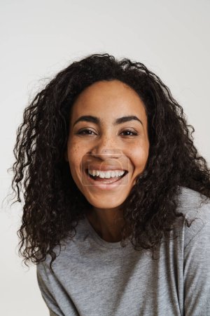 Téléchargez les photos : Jeune femme noire aux cheveux ondulés riant et regardant la caméra isolée sur fond blanc - en image libre de droit