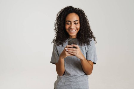 Téléchargez les photos : Jeune femme noire utilisant un téléphone portable et riant isolé sur fond blanc - en image libre de droit