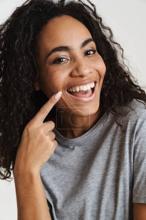 Foto de Joven mujer negra con el pelo ondulado señalando con el dedo su sonrisa aislada sobre fondo blanco - Imagen libre de derechos