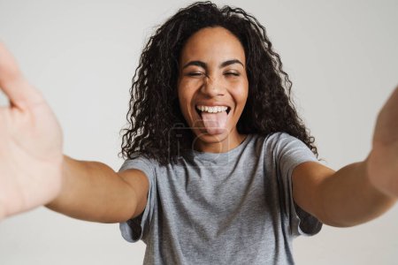 Foto de Joven mujer negra mostrando su lengua mientras toma foto autofoto aislada sobre fondo blanco - Imagen libre de derechos
