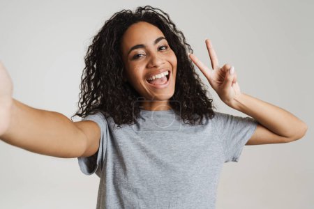 Photo for Young black woman showing peace sign while taking selfie photo isolated over white background - Royalty Free Image