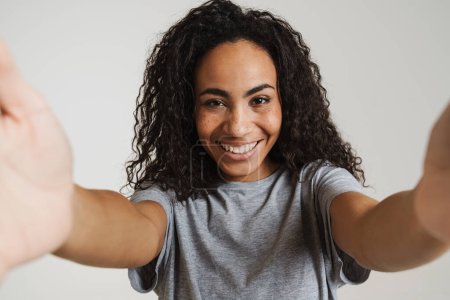 Foto de Joven mujer negra con el pelo ondulado sonriendo mientras toma foto autofoto aislada sobre fondo blanco - Imagen libre de derechos