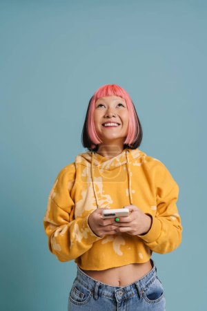 Foto de Asiática chica con el pelo rosa sonriendo y utilizando el teléfono móvil aislado sobre fondo azul - Imagen libre de derechos