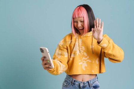 Photo for Asian girl with pink hair gesturing while taking selfie on cellphone isolated over blue background - Royalty Free Image
