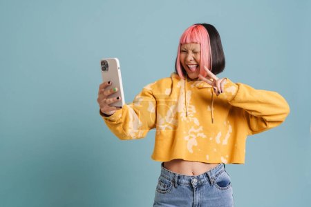Téléchargez les photos : Asiatique fille avec rose cheveux geste tout en prenant selfie sur téléphone portable isolé sur fond bleu - en image libre de droit