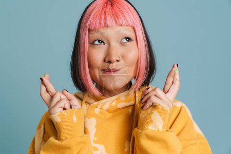 Téléchargez les photos : Asiatique fille avec les cheveux roses tenant les doigts croisés pour bonne chance isolé sur fond bleu - en image libre de droit