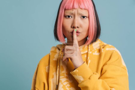 Téléchargez les photos : Asiatique fille avec les cheveux roses et piercing montrant geste de silence isolé sur fond bleu - en image libre de droit