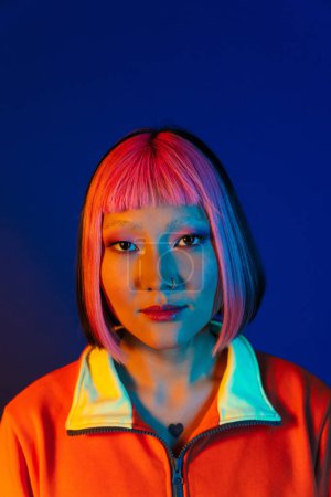 Téléchargez les photos : Asiatique fille avec des cheveux roses et piercing posant tout en regardant la caméra isolée sur fond bleu - en image libre de droit