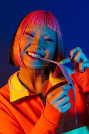 Téléchargez les photos : Asiatique fille avec rose cheveux faire plaisir tout en mangeant mastication marmelade isolé sur fond bleu - en image libre de droit