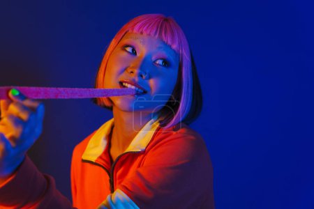 Photo for Asian girl with pink hair making fun while eating chewing marmalade isolated over blue background - Royalty Free Image
