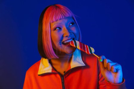 Foto de Chica asiática con el pelo rosa burlándose mientras come helado aislado sobre fondo azul - Imagen libre de derechos