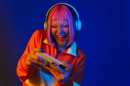 Téléchargez les photos : Asiatique fille dans casque jouer jeu en ligne sur téléphone mobile isolé sur fond bleu - en image libre de droit