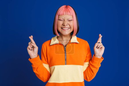 Téléchargez les photos : Asiatique fille avec rose cheveux rire tout en tenant les doigts croisés isolé sur fond bleu - en image libre de droit