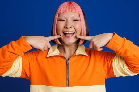 Foto de Asiática chica con el pelo rosa y piercing señalando los dedos a su sonrisa aislado sobre fondo azul - Imagen libre de derechos