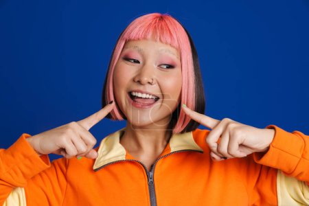 Foto de Asiática chica con el pelo rosa y piercing señalando los dedos a su sonrisa aislado sobre fondo azul - Imagen libre de derechos