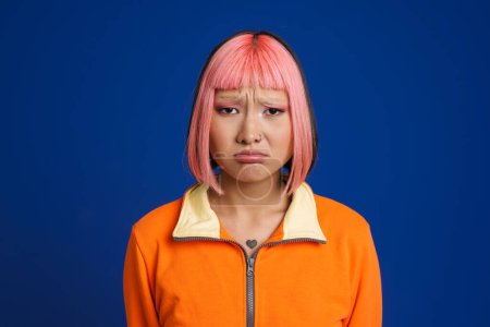 Téléchargez les photos : Asiatique fille avec des cheveux roses et perçants fronçant les sourcils tout en regardant la caméra isolée sur fond bleu - en image libre de droit