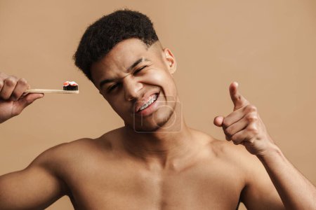 Foto de Joven hombre sin camisa apuntando con el dedo a la cámara mientras posa con cepillo de dientes aislado sobre fondo beige - Imagen libre de derechos