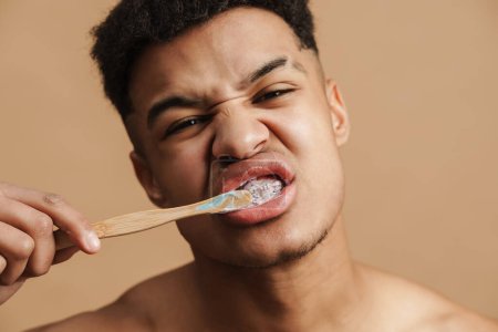 Jeune homme torse nu fronçant les sourcils tout en se brossant les dents isolé sur fond beige