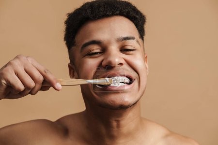 Jeune homme torse nu souriant tout en se brossant les dents isolé sur fond beige