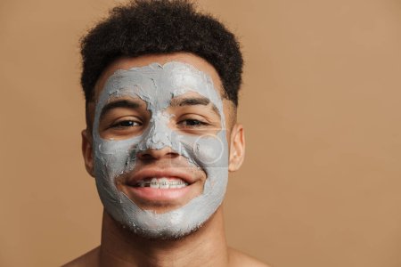 Foto de Joven hombre sin camisa con máscara de arcilla cosmética sonriendo a la cámara aislada sobre fondo beige - Imagen libre de derechos