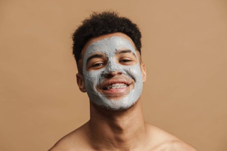 Foto de Joven hombre sin camisa con máscara de arcilla cosmética sonriendo a la cámara aislada sobre fondo beige - Imagen libre de derechos