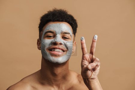 Foto de Hombre sin camisa con máscara de arcilla cosmética sonriendo y haciendo gestos a la cámara aislada sobre fondo beige - Imagen libre de derechos