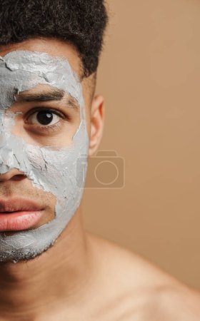 Téléchargez les photos : Jeune homme torse nu avec masque en argile cosmétique regardant la caméra isolée sur fond beige - en image libre de droit