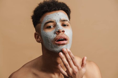 Téléchargez les photos : Jeune homme torse nu avec masque en argile cosmétique touchant son menton isolé sur fond beige - en image libre de droit