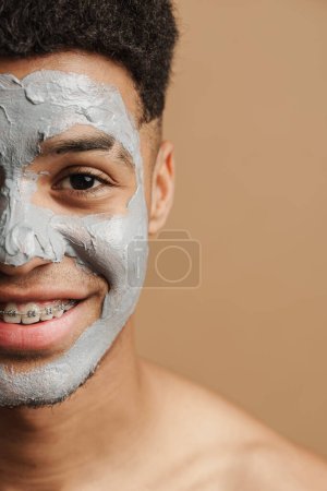 Téléchargez les photos : Jeune homme torse nu avec masque en argile cosmétique souriant à la caméra isolé sur fond beige - en image libre de droit