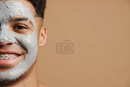 Téléchargez les photos : Jeune homme torse nu avec masque en argile cosmétique souriant à la caméra isolé sur fond beige - en image libre de droit