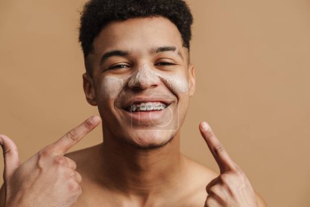 Téléchargez les photos : Jeune homme torse nu avec masque d'argile cosmétique pointant du doigt son visage isolé sur fond beige - en image libre de droit
