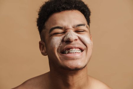 Photo for Young shirtless man with cosmetic clay mask smiling at camera isolated over beige background - Royalty Free Image