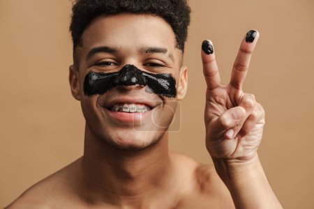Téléchargez les photos : Homme torse nu avec masque cosmétique souriant et gestuel à la caméra isolé sur fond beige - en image libre de droit