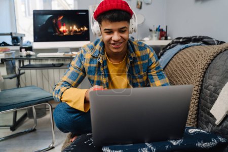 Photo for Middle-eastern teenage boy using laptop and cellphone while sitting on couch at home - Royalty Free Image