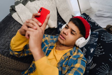 Téléchargez les photos : Moyen-Orient adolescent garçon à l'aide d'écouteurs et téléphone portable tout en étant couché sur le canapé à la maison - en image libre de droit