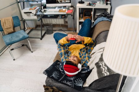 Téléchargez les photos : Moyen-Orient adolescent garçon à l'aide d'écouteurs et téléphone portable tout en étant couché sur le canapé à la maison - en image libre de droit