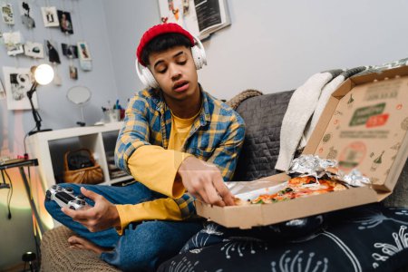 Téléchargez les photos : Moyen-Orient adolescent garçon manger pizza et en utilisant contrôleur tout en jouant jeu vidéo à la maison - en image libre de droit