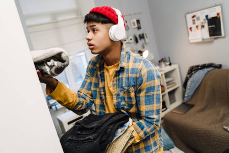 Photo for Teenage boy using headphones folding his clothes while cleaning bedroom at home - Royalty Free Image