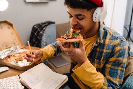 Téléchargez les photos : Moyen-Orient adolescent garçon manger pizza tout en étudiant à la maison par ordinateur - en image libre de droit
