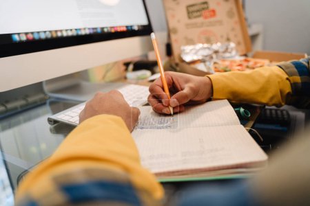 Téléchargez les photos : Adolescent garçon écriture dans cahier d'exercices et en utilisant ordinateur de bureau tout en étudiant à la maison - en image libre de droit
