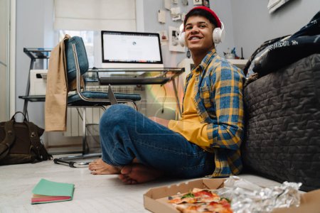 Téléchargez les photos : Moyen-Orient adolescent garçon en utilisant un ordinateur portable et manger de la pizza tout en étudiant à la maison - en image libre de droit