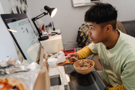 Téléchargez les photos : Brunette adolescent garçon à l'aide d'un ordinateur de bureau tout en prenant le petit déjeuner dans la chambre - en image libre de droit
