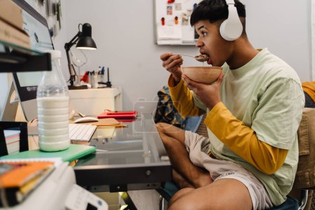 Téléchargez les photos : Brunette adolescent garçon à l'aide d'un ordinateur de bureau tout en prenant le petit déjeuner dans la chambre - en image libre de droit