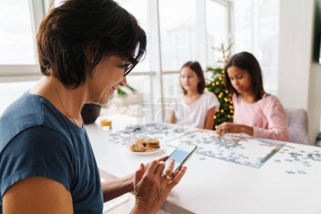 Téléchargez les photos : Sœurs multiraciales faisant puzzle ensemble tandis que leur mère utilisant un téléphone portable à la maison - en image libre de droit