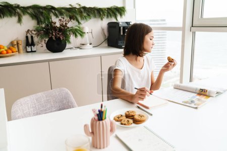 Photo for White preteen girl eating cookie while doing homework at home - Royalty Free Image