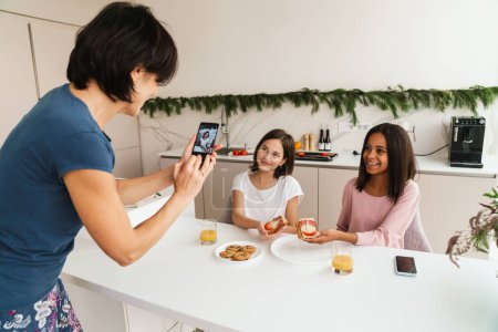 Téléchargez les photos : Femme blanche souriant tout en prenant des photos de ses filles dans la cuisine à la maison - en image libre de droit