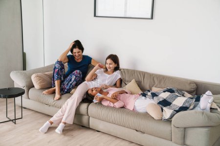 Téléchargez les photos : Femme blanche et ses filles riant tout en se reposant sur le canapé à la maison - en image libre de droit