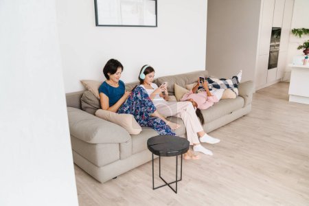 Téléchargez les photos : Femme blanche et ses filles en utilisant des gadgets tout en se reposant sur le canapé à la maison - en image libre de droit