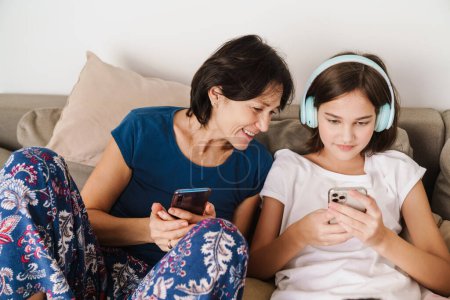 Téléchargez les photos : Femme blanche et sa fille en utilisant des gadgets tout en se reposant sur le canapé à la maison - en image libre de droit
