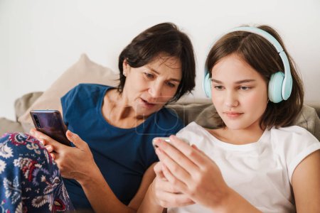 Téléchargez les photos : Femme blanche et sa fille en utilisant des gadgets tout en se reposant sur le canapé à la maison - en image libre de droit